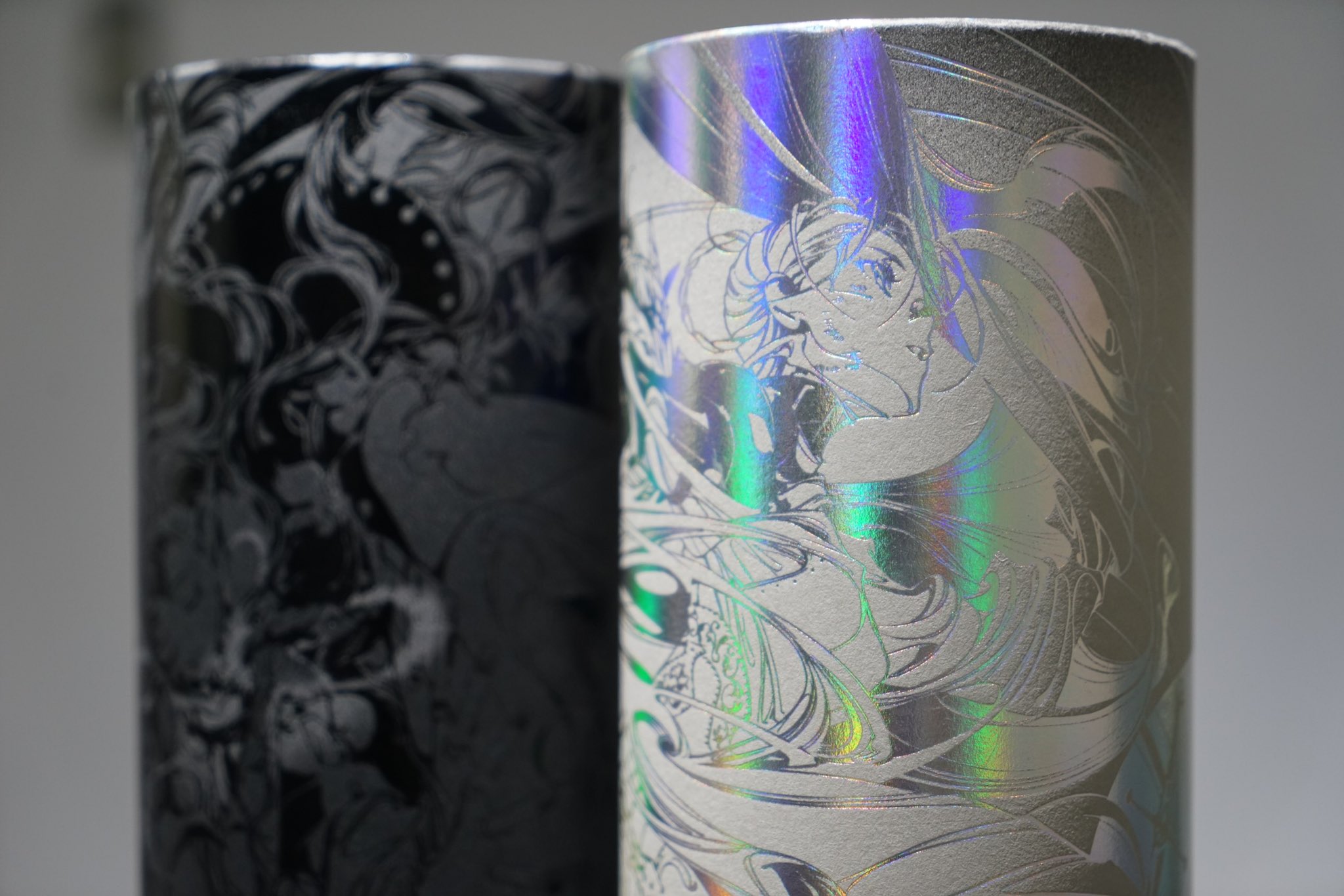 two cylindrical containers are shown sidebyside. the container on the left features a blackandwhite illustration of a woman with long flowing hair. the container on the right features a similar illustration, but it is printed on a holographic material that shimmers with rainbow colors. the containers are lit from the side, creating a sense of depth and dimension.