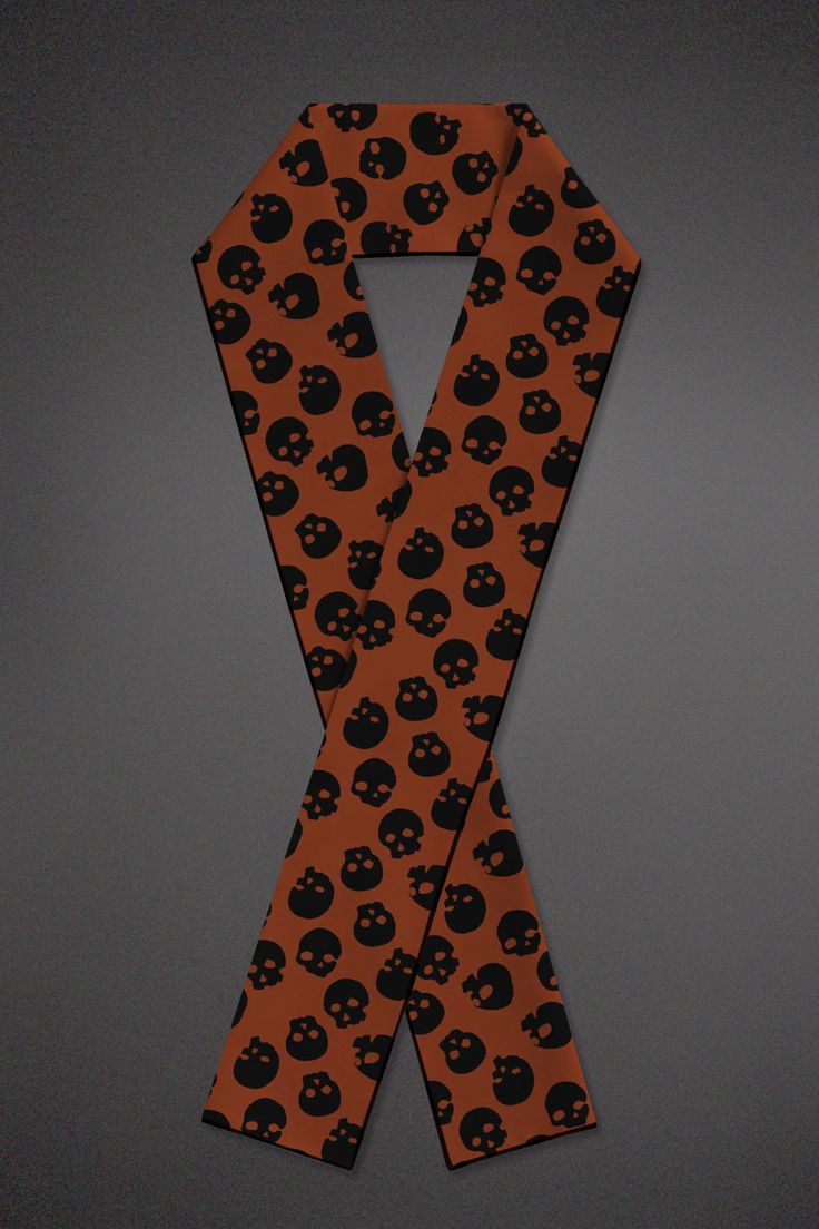 an orange scarf with a black skull pattern is displayed against a dark gray background. the scarf is arranged in a loose, crossed manner, showcasing the repeating skull design.