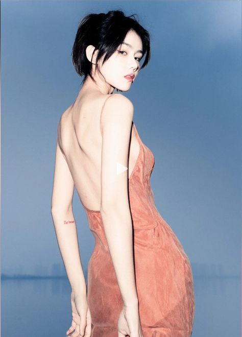 a woman with short dark hair wearing a lowback dress looks over her shoulder at the camera. she is standing outdoors in front of a body of water and the sky. she has a small tattoo on her left arm.