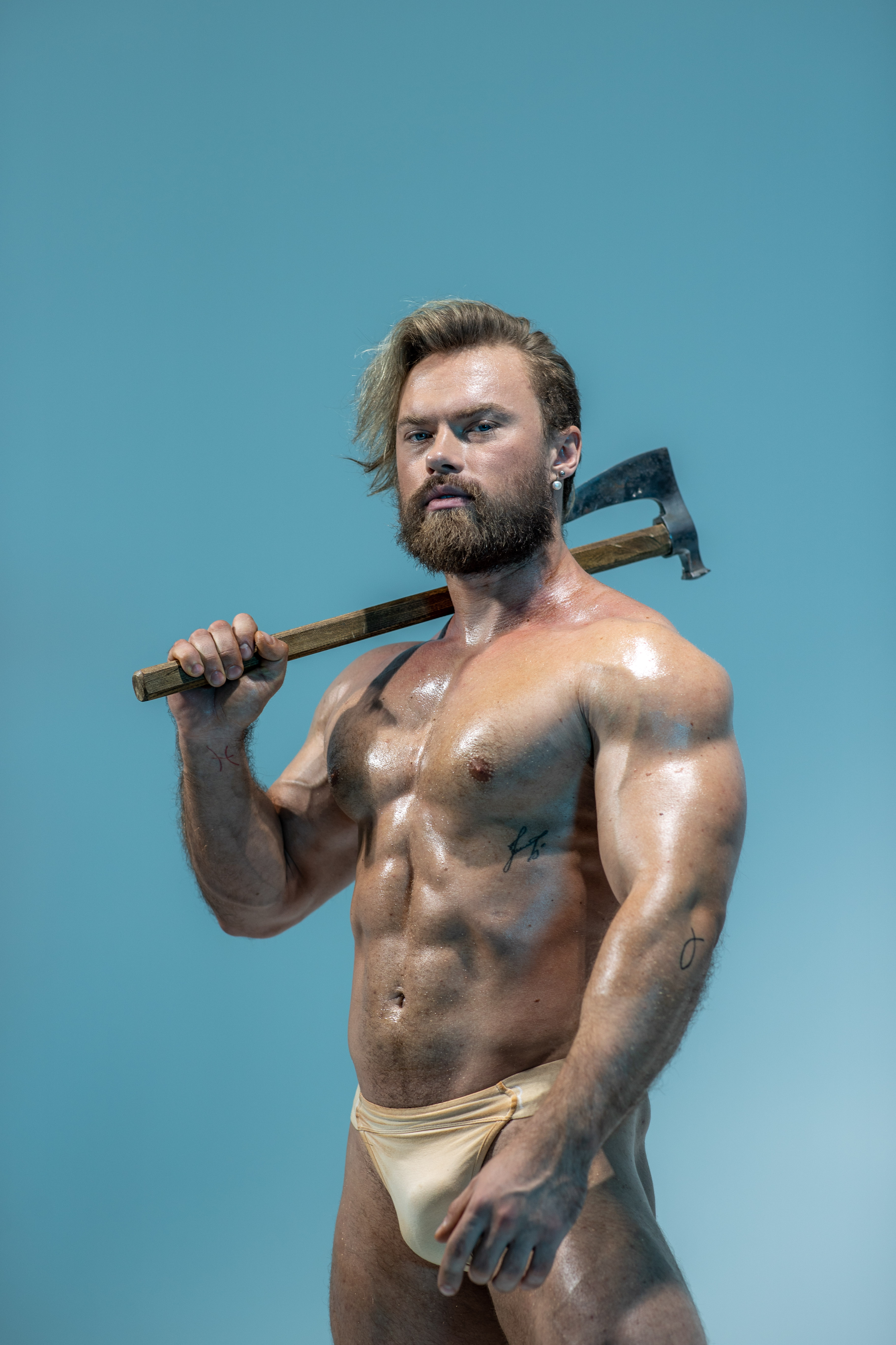 a muscular, blonde man with a beard and long hair stands in front of a blue backdrop. he's wearing only beige underwear and holds a worn axe across his shoulders, gripping it with both hands. his body is glistening, suggesting oil or sweat.  he stares intently at the viewer, conveying an aura of power and masculinity.