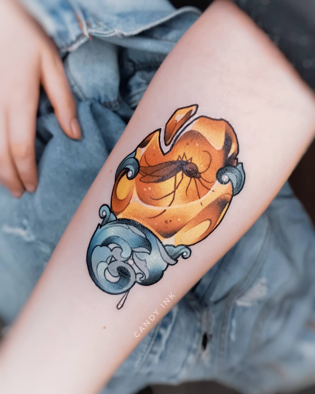 a tattoo of a mosquito trapped in amber on a forearm. the amber piece has a decorative blue base with wave patterns. the tattoo is done in a colorful illustrative style with black outlines.