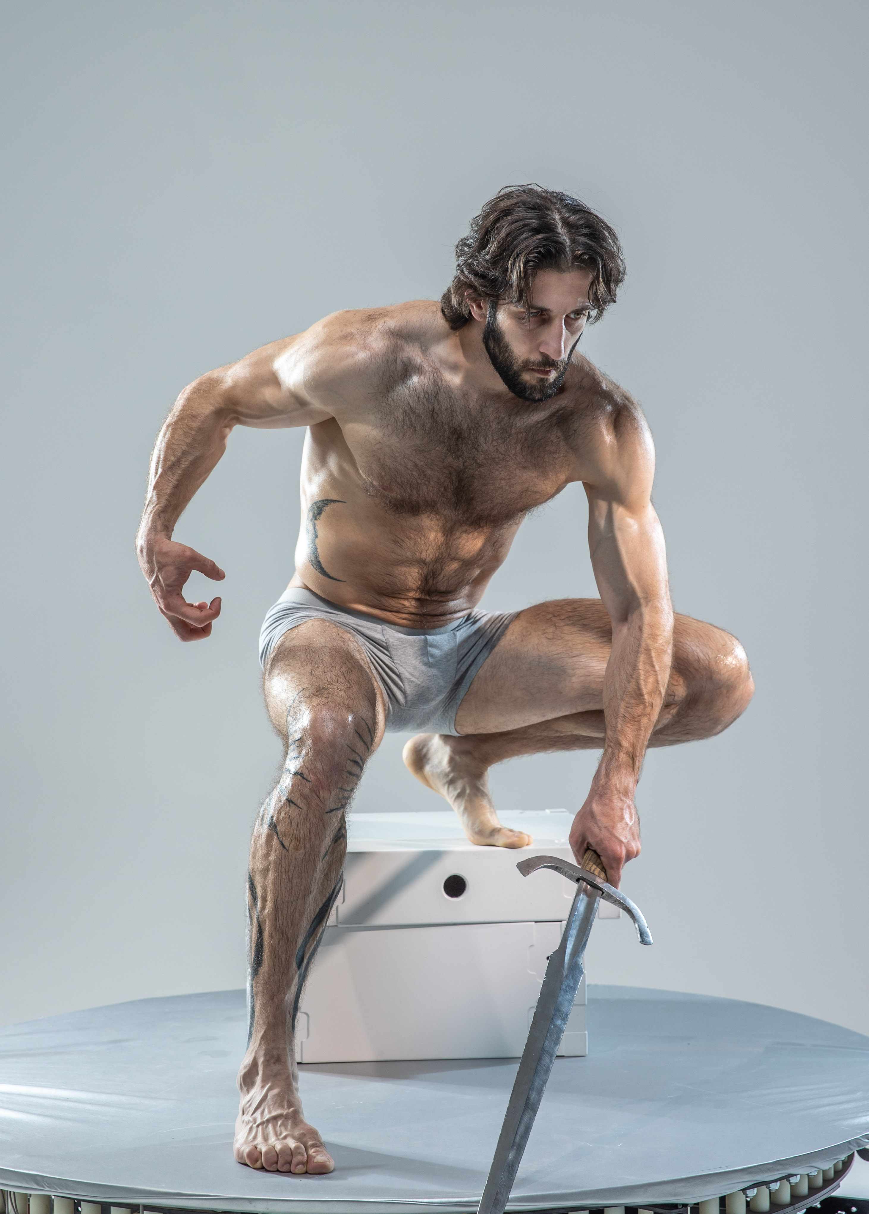 a muscular man with a beard and long hair is posing with a sword. he is wearing only underwear and has a tattoo on his leg. he is standing on a white box on a rotating platform in a studio. he is lit with soft light from above. the perspective is low angle.