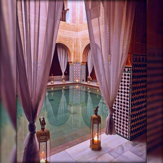 a photo of an intricately tiled indoor pool.  the tranquil scene is softly lit by lanterns and candles, and sheer curtains add to the luxurious and serene atmosphere. the mosaic tiles create a vibrant backdrop while the water in the pool reflects the light, adding depth and ambiance.