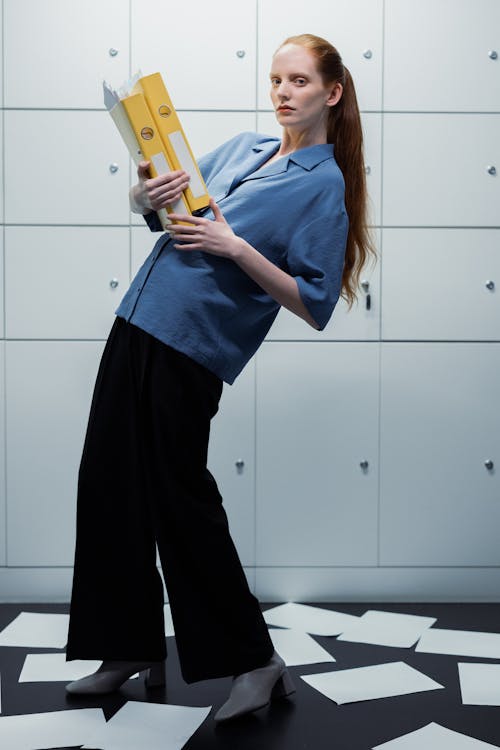 a full shot of a woman with long red hair tied back in a ponytail, wearing a light blue buttonup shirt and black trousers, carrying two yellow files while leaning slightly to her left. she's standing on a dark floor amid scattered white papers in a minimalist room with lightgrey wall cabinets.  she has a serious expression. the overall style is stylish and modern, suggestive of a professional, business setting.