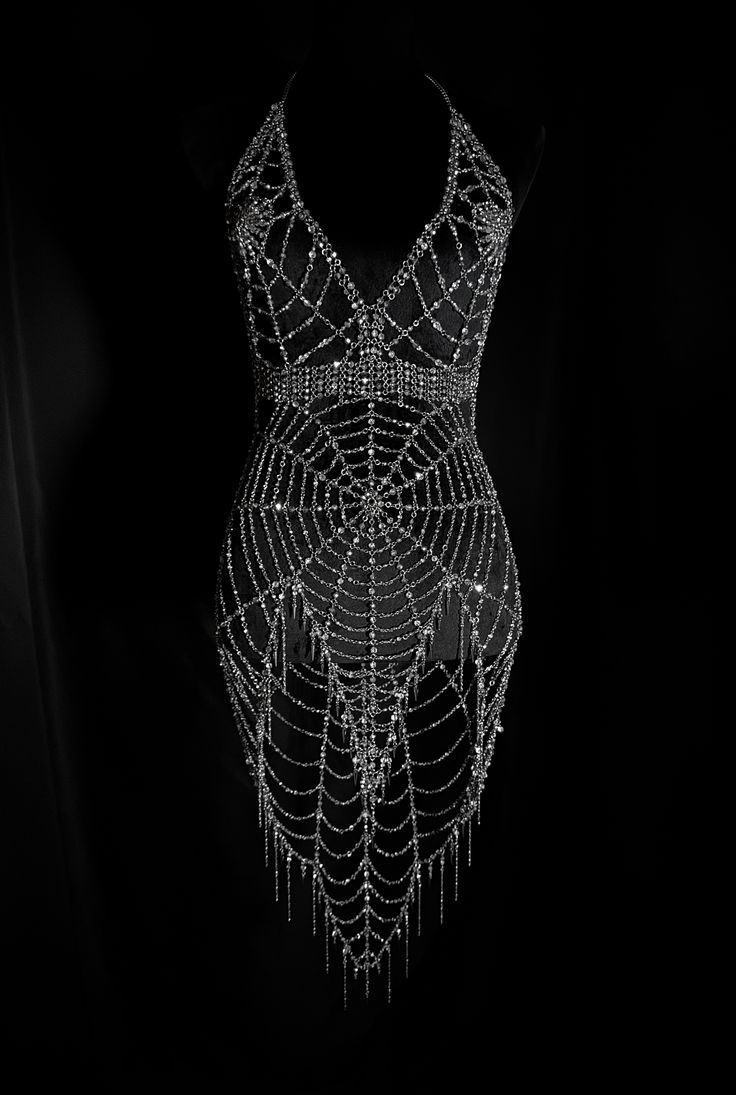 a black and white photograph showcases a dress adorned with intricate spiderweb patterns. the dress features a halter neck design and is heavily embellished with shiny beads. the spiderweb patterns cascade down the dress, creating a visually striking effect. hanging beads further enhance the dress's elegance and texture. the dark background accentuates the dress, making it the focal point of the image. the garment appears to be evening or formal wear, designed for special occasions.