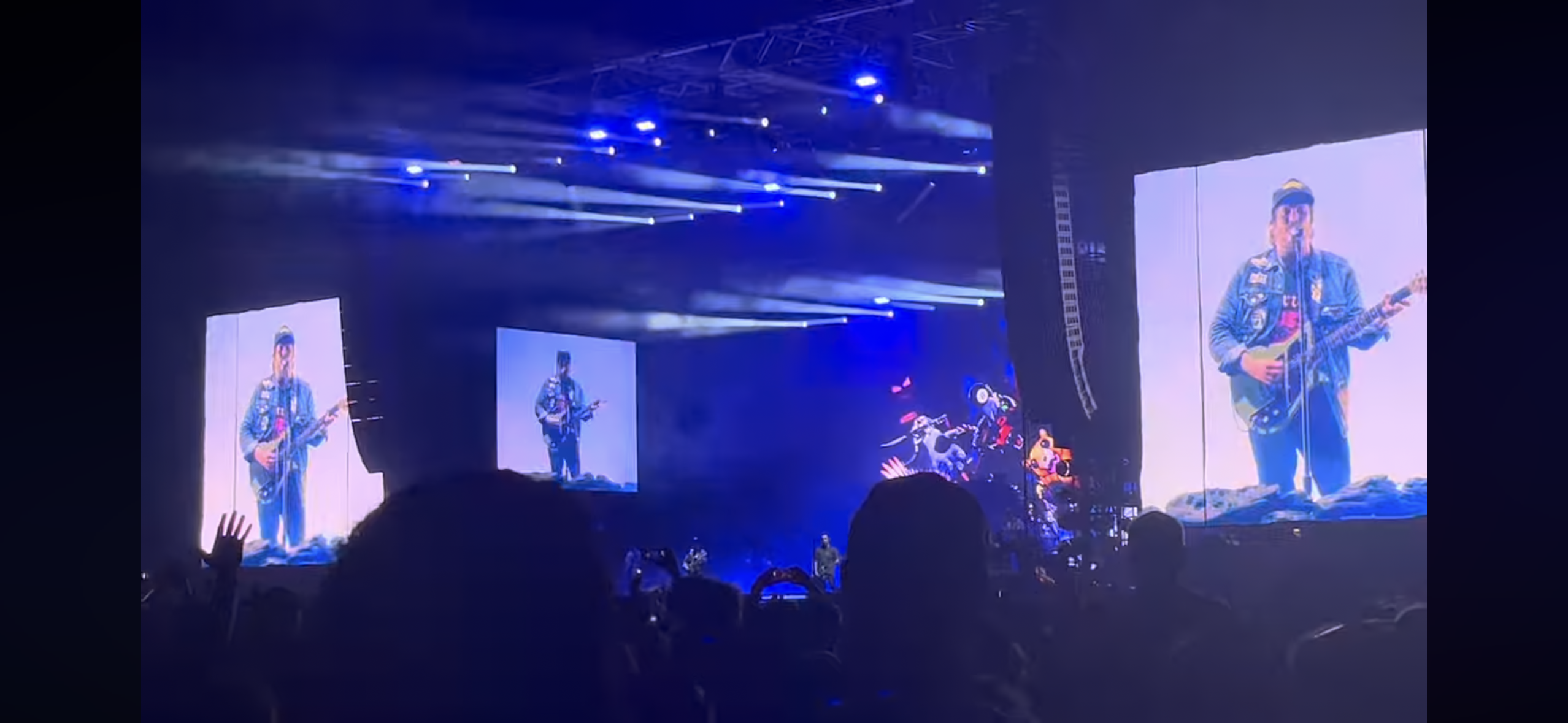 a concert scene captured from the audience perspective, showcasing a band on stage with their lead singer in the spotlight.  the singer is playing an electric guitar and is singing passionately. behind him, are multiple large screens displaying the singer’s image and the band's music. the stage is illuminated by dramatic lighting effects, creating a vibrant atmosphere. the crowd can be seen in the foreground, excitedly cheering and enjoying the music. the energy of the concert is palpable, with the music, the lights, and the crowd all coming together to create a memorable experience.