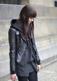 a young woman with long brunette hair with bangs is wearing a black leather jacket with a hood, black skirt, and black pants. she is looking down at the ground with a serious expression. the woman is standing in the city.