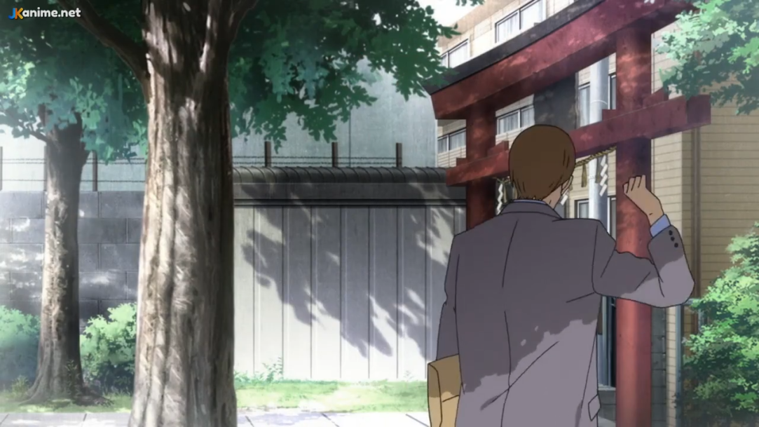 a man in a white shirt and gray suit jacket walks away from the camera down a sidewalk towards a torii gate. he is holding a brown paper bag in his left hand and waving with his right hand. there is a large green tree on the left side of the image and a gray fence runs behind it. the building behind the torii gate is beige with brown trim.  the sun in the sky casts shadows from the fence and tree.