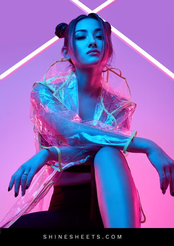 a young woman in a transparent raincoat is sitting and posing for a photoshoot. she is illuminated by neon lights in pink and blue, giving her a futuristic and stylish look. her hair is styled in buns and her expression is serious.