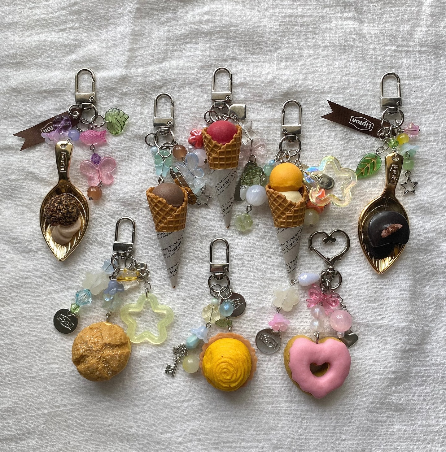 the image displays several keychains with dessertthemed charms. some feature ice cream cones with different flavored scoops, while others showcase chocolate candies on golden spoons. additionally, there's a pink donut, a yellow pastry, and various colorful beads and charms adorning each keychain. a ribbon with the word "lipton" is also visible on one of the keychains.