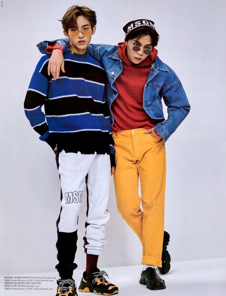 the photo shows two young asian men posing in front of a white background. they are both wearing trendy and casual clothes. the man on the left is wearing a blue and black striped sweater, white and black pants, and black and white sneakers. he is also wearing sunglasses. the man on the right is wearing a denim jacket over a red hoodie, yellow pants, and black shoes. he is also wearing a beanie. they are looking at the camera, and the man on the left has his arm around the man on the right. the photo captures a moment of friendship and style.