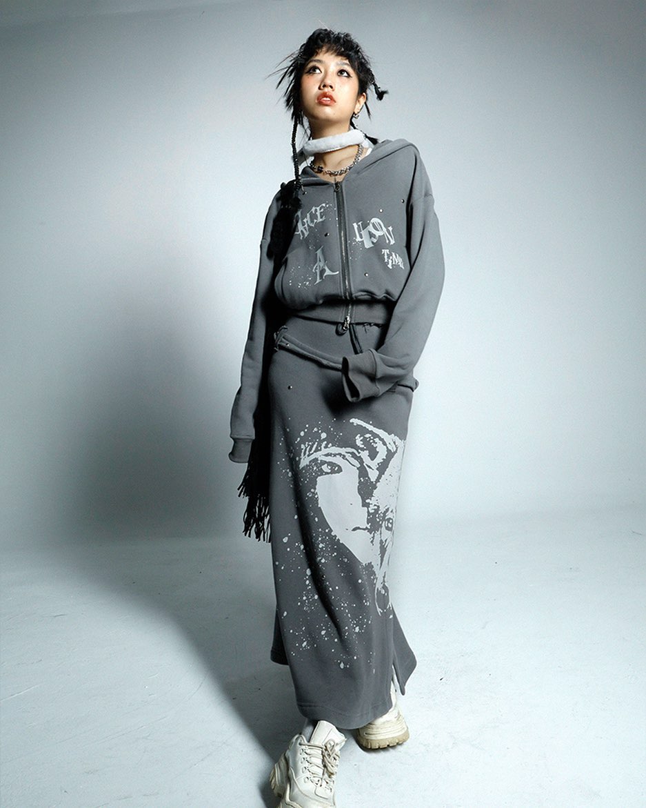 a young woman model is posing in a grey hoodie and matching long skirt set. the set features a graphic print on the skirt, and the hoodie has text print. the model is wearing white sneakers and has her hair styled in braids. she is looking up and to the side, with a slightly pensive expression. the background is a plain, light grey studio setting.
