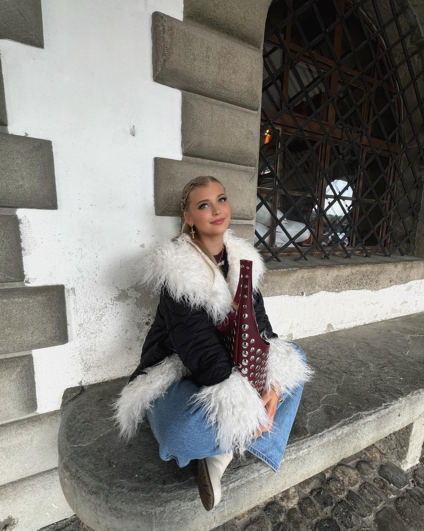 a young woman with blonde braided hair is sitting on a stone ledge in front of an architectural building. she is wearing a black jacket with white fur trim, light blue jeans, and white boots. she holds a maroon handbag adorned with metal studs. the background features a stone wall with distinct patterns and a barred window. she looks upward with a slight smile.