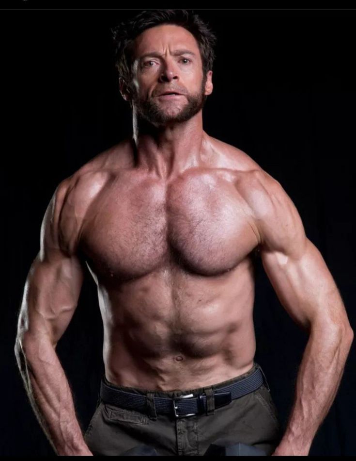 a man with a muscular physique stands shirtless in front of a black background. his arms are flexed slightly, showcasing his biceps and triceps.  he has a beard and stubble, and is looking directly at the camera. he is wearing a belt around his waist, but the rest of his torso is bare, revealing a sixpack and welldefined chest. the man appears to be in excellent physical condition, with strong muscles throughout his body.