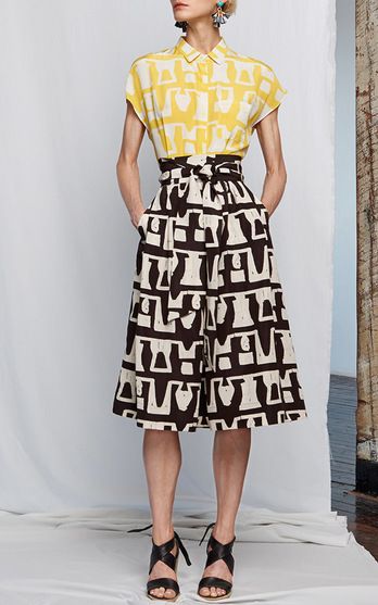 a fullbody shot of a fashion model showcasing a stylish outfit. the model, a woman with fair skin, is wearing a yellow shortsleeved shirt with a geometric print and a black and white midi skirt with a matching pattern. the skirt has a defined waistline with a belt. she accessorizes with black strappy sandals and dangling earrings.  the background is simple, a white studio setting with a hint of wood flooring visible in the lower corner. the overall image is clean and focuses on the model and her clothing.