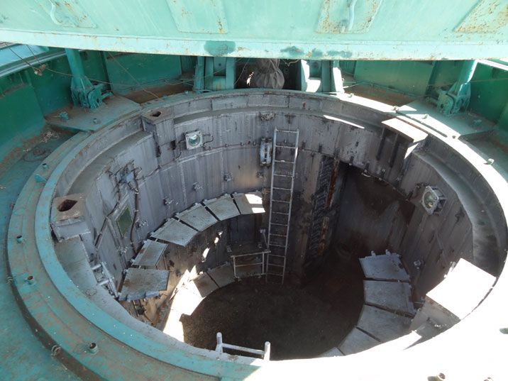 the image shows the interior of an abandoned, likely missile, silo. a large, circular opening is visible, revealing the silo's depth and internal structure. the walls are made of aged, grey concrete and metal. a metal ladder descends into the darkness below. several metal plates are arranged in a spiral pattern on the silo's walls, possibly remnants of a mechanism or support structures. the top of the silo features a large, partially open, teal green hatch. the overall impression is one of dereliction and age, suggesting a site from a bygone era, possibly related to the cold war. the lighting indicates it is daytime, with sunlight partially illuminating the silo's interior.