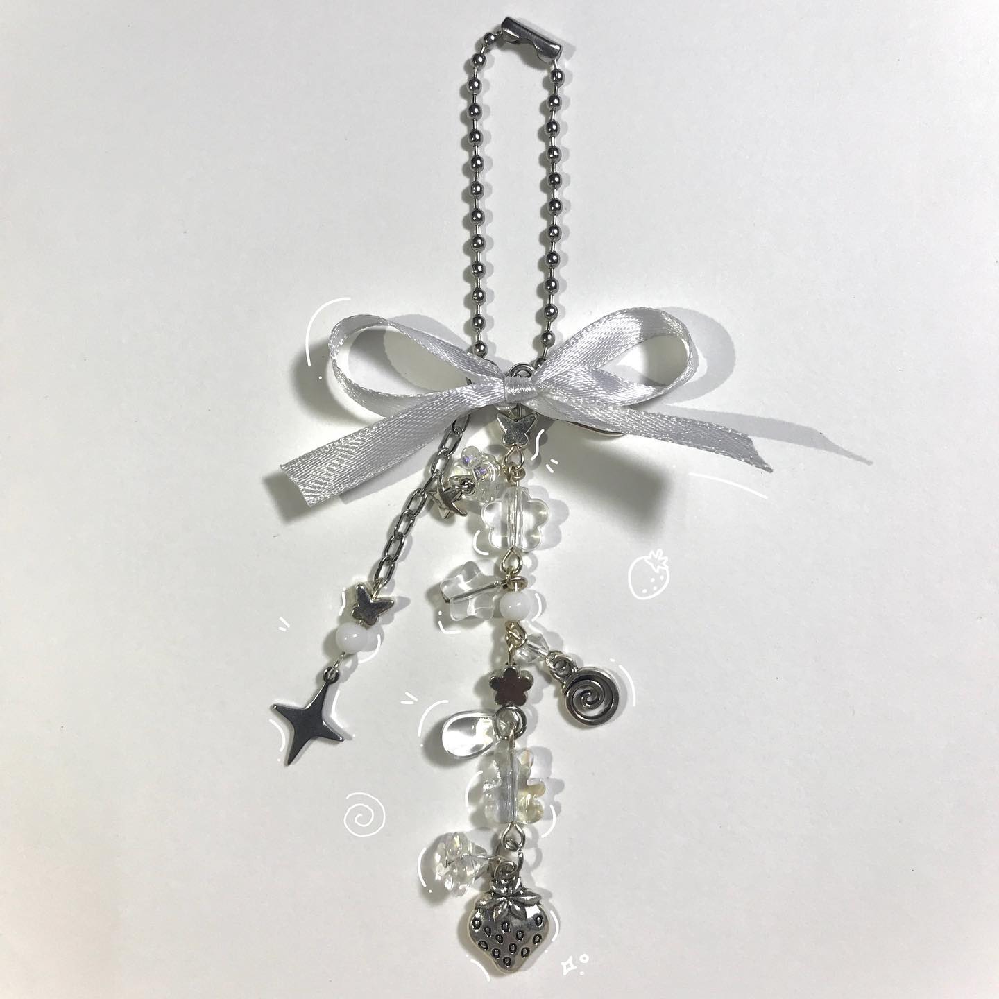 a closeup shot of a handmade keychain. it features a silver ball chain with a clip at the top, a tied silver ribbon, and a variety of beads and charms hanging below. the beads are mostly clear or white, and the charms include a star, a flower, a spiral, and a strawberry. the keychain is on a white surface.