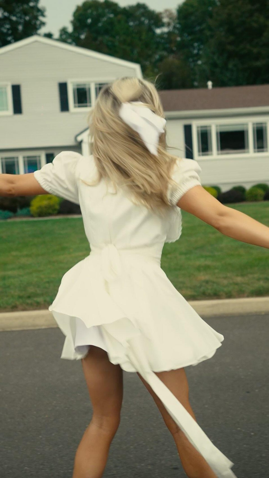 a woman is seen from the back, wearing a short, white dress and a matching ribbon in her blonde hair, while standing on a road in a suburban neighborhood. she has her arms outstretched to the sides. in the background are houses with green lawns and trees. the sky appears overcast, suggesting it is daytime but not sunny.
