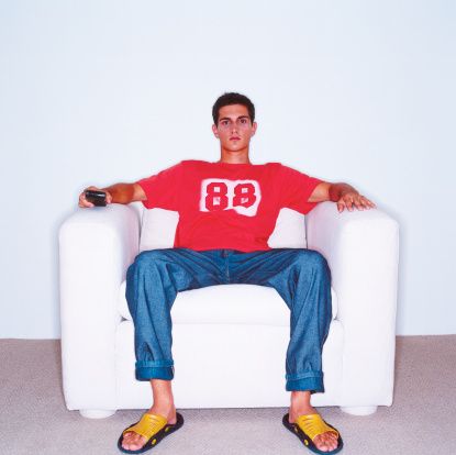 a man is sitting on a white couch, relaxing and watching television. he is wearing jeans and a red shirt with a white number 88 on it. he is holding a remote control in his hand. he looks comfortable and relaxed.