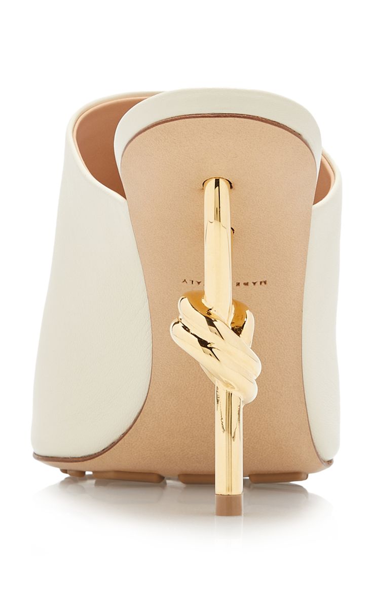 a closeup back view of a pair of by far white leather mules with a distinctive gold knot heel. the shoes are elegant and stylish, perfect for a variety of occasions.  the image showcases the detail of the heel and the overall design of the shoe.