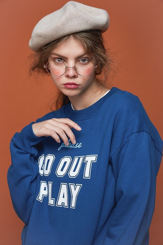 a young woman poses in a studio shot against a burnt orange background. she is wearing a beige beret, round glasses, and a royal blue sweatshirt with the words "please go out play" printed on the front. her hair is loose, and she has a slightly serious expression. the overall style is casual and stylish.