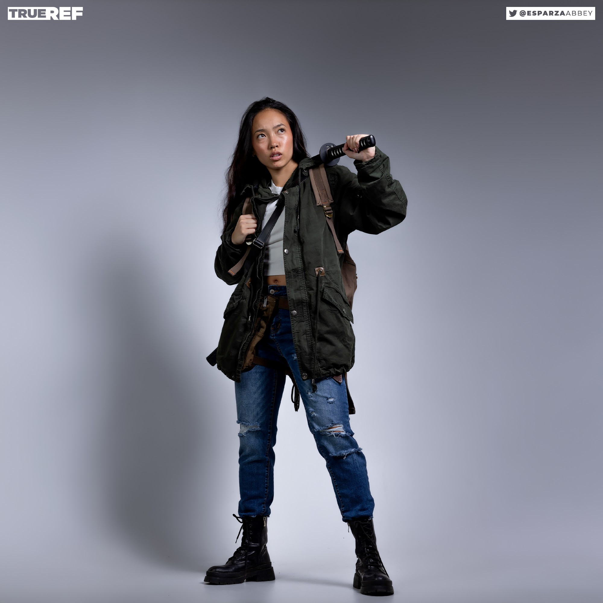 a fullbody shot of a young asian woman, appearing determined, stands against a light gray backdrop. she's dressed in a dark olive green parka jacket, distressed blue jeans, and combat boots.  a backpack is slung over her shoulders. she holds a katana (a type of japanese sword) aloft, looking upward.  the overall style suggests a postapocalyptic or survivalist theme.  the image is welllit and professionally photographed, possibly for a fashion or modeling portfolio.