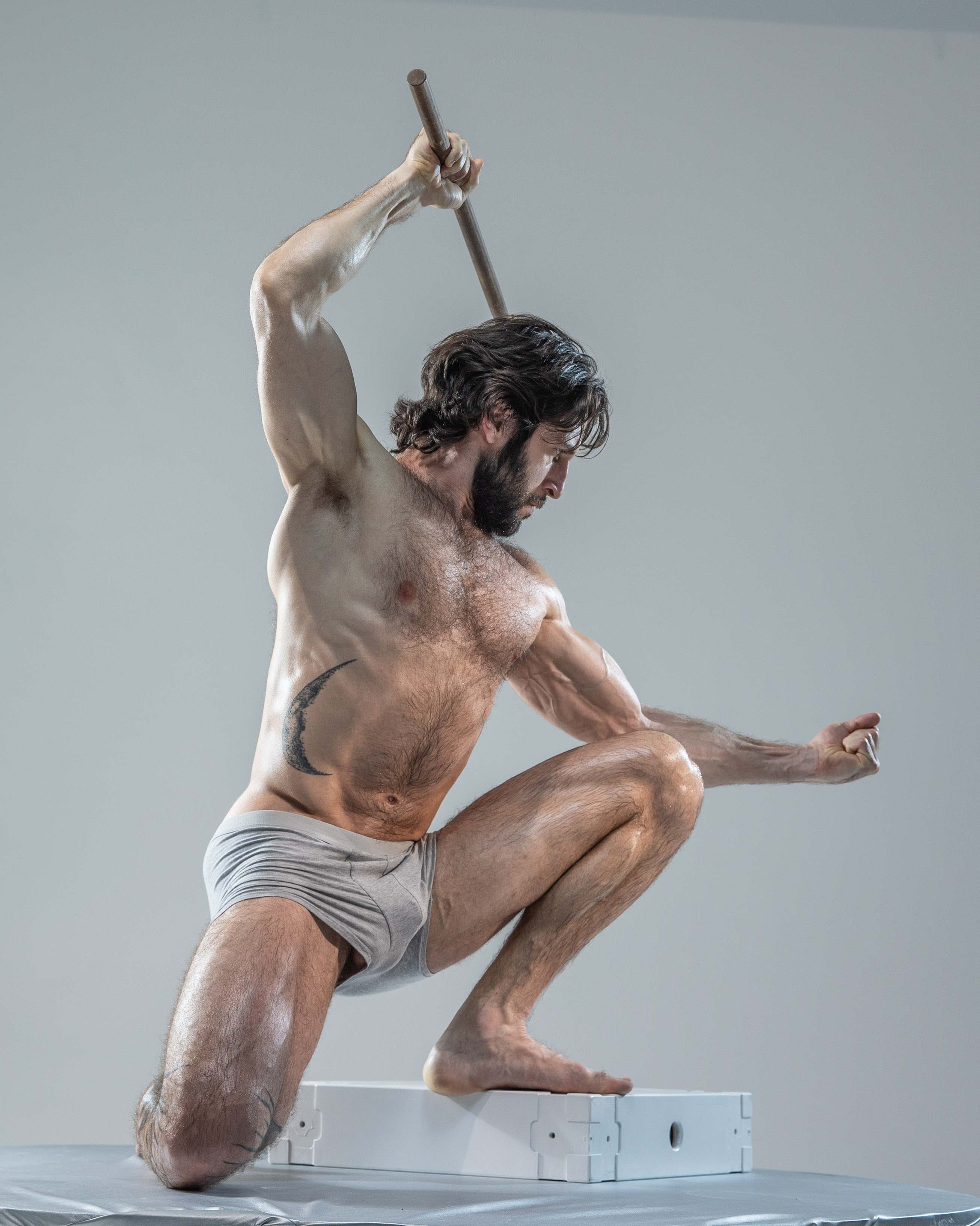 a muscular, hairy man in grey underwear poses in a dynamic fighting stance against a plain, light grey studio background. he holds a wooden staff above his head, as if about to strike, and his body is tensed with energy.  the low camera angle emphasizes his strength and power.