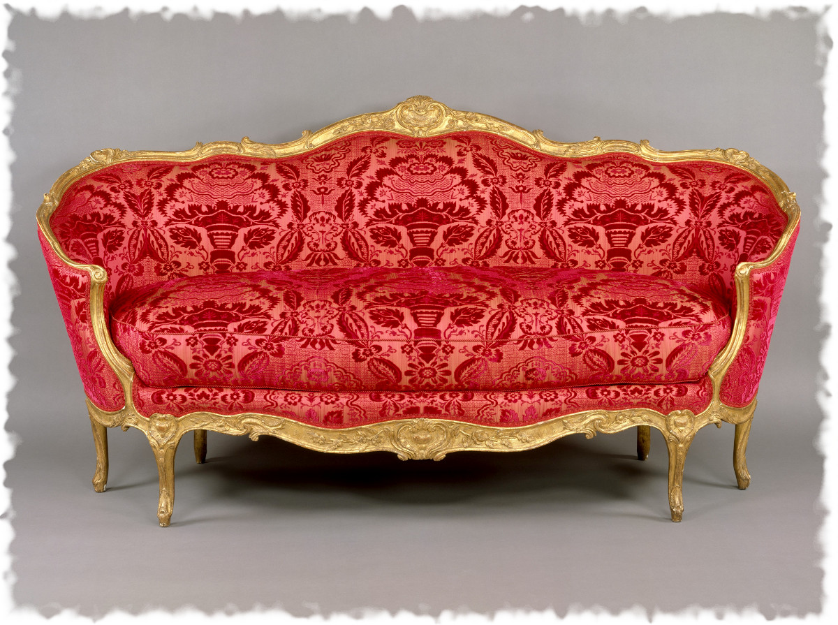 a rococostyle sofa with a red patterned velvet upholstery and ornate gilded gold frame stands centered against a neutral gray background. the sofa features a curved back and seat, with intricate carvings along the frame and legs. the fabric is adorned with a floral and geometric pattern. the overall impression is one of opulence and vintage luxury.
