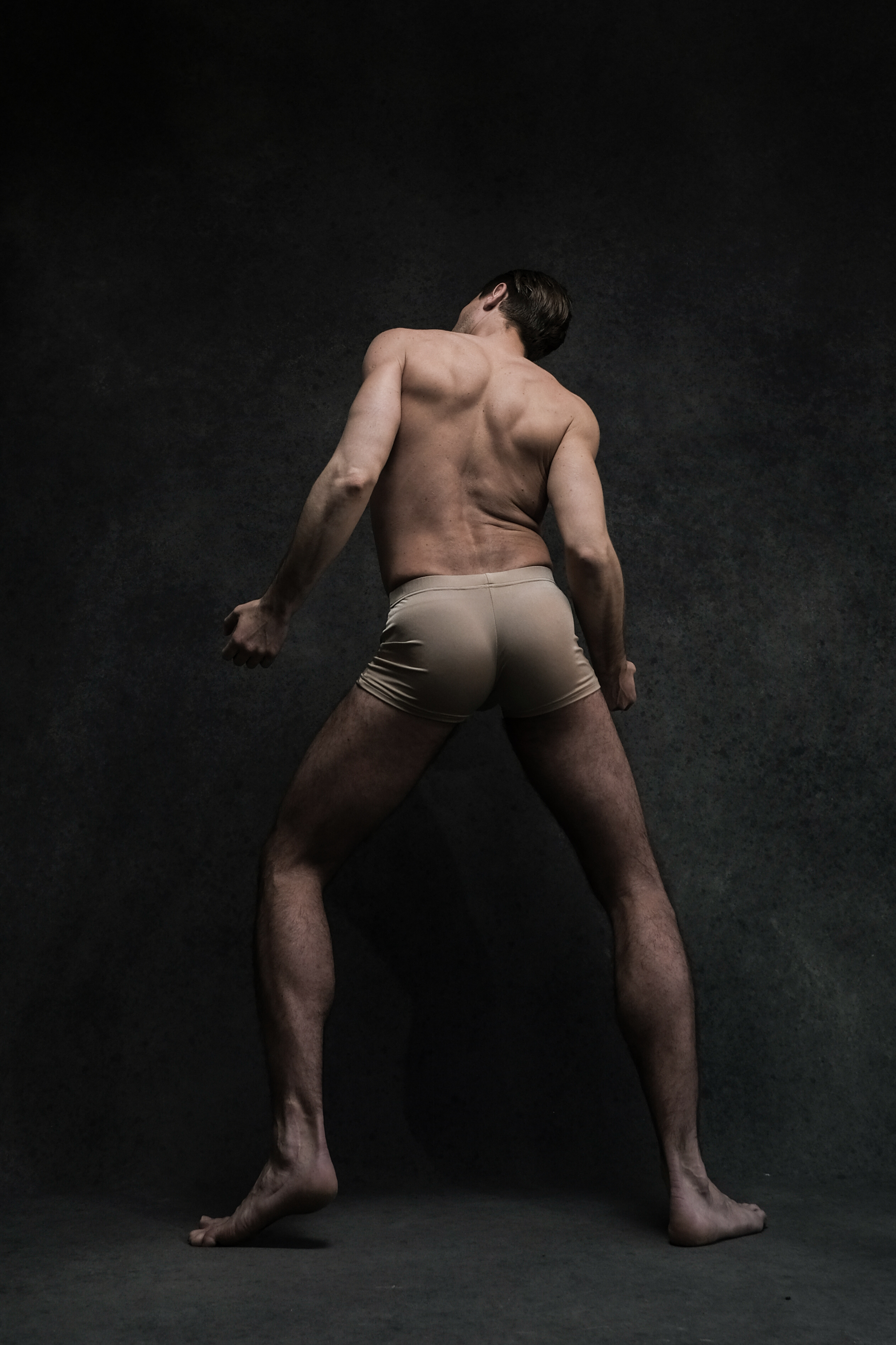 a male dancer, wearing only tan shorts, is captured midpose against a dark, textured background. the dramatic lighting emphasizes the musculature of his back, legs, and arms, highlighting the strength and flexibility required for dance. his posture suggests a dynamic movement, frozen in time by the photograph.