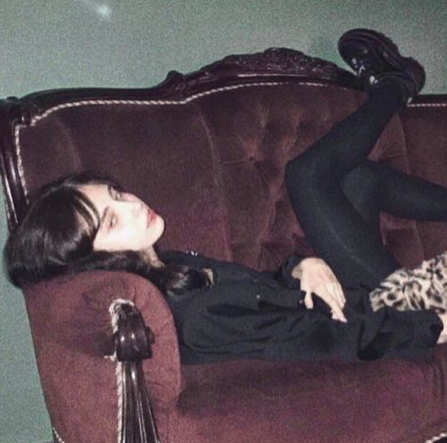 a woman is lying on a burgundy velvet couch with her legs up. she is wearing black clothes and black shoes. there is a leopard print blanket on the couch next to her. the background is a green wall.