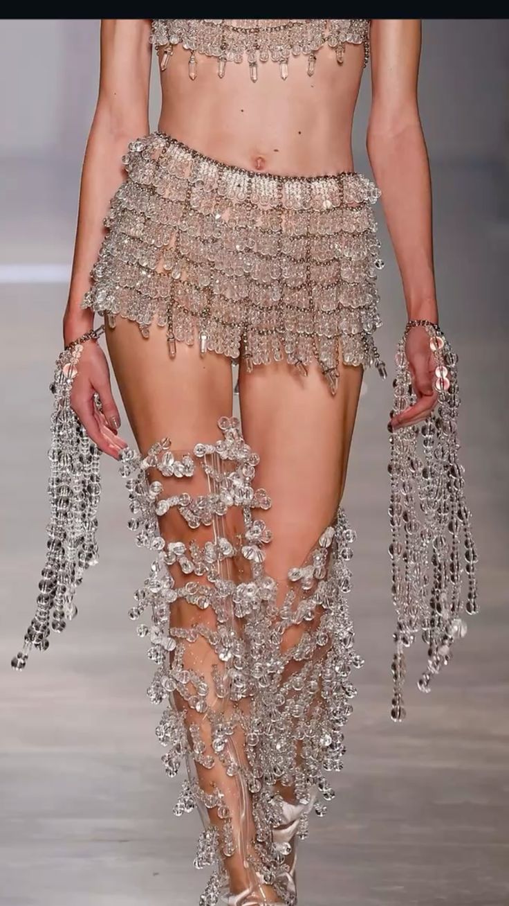a model walks the runway wearing a striking twopiece outfit heavily embellished with clear beads and crystals. the outfit includes a bra top and a short, tiered skirt, both entirely covered in shimmering beads. clear, kneehigh boots adorned with more crystals and long, beaded gloves complete the look. the model's bare midriff is visible between the top and skirt. the setting appears to be a fashion show, with a neutralcolored runway visible in the background.