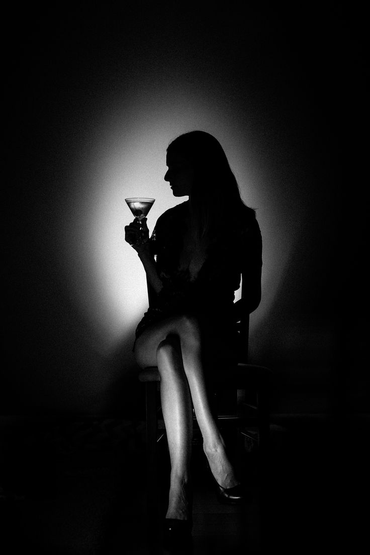 a woman in a silhouette is sitting in a chair holding a martini glass in a dark room with a spotlight on her. she is dressed elegantly, and her legs are crossed. the image has a moody, mysterious, and seductive feel.
