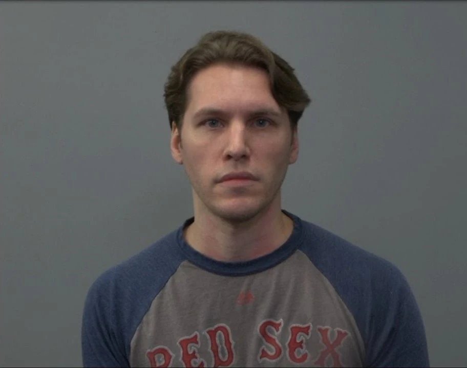 a man with short, light brown hair is pictured in a headshot against a solid gray background. he has a neutral expression on his face. he's wearing a blue and gray baseballstyle tshirt.