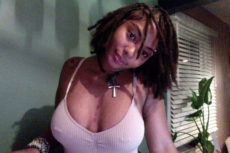 a closeup selfie of a young african american woman with dreadlocks. she's wearing a light pink camisole, a necklace with a cross pendant, and has several visible body piercings including a septum piercing and nipple piercings.  there are also tattoos on her shoulder and chest. she appears relaxed, and the background shows a bedroom setting with a window and blinds, and a houseplant. the lighting is soft and natural.