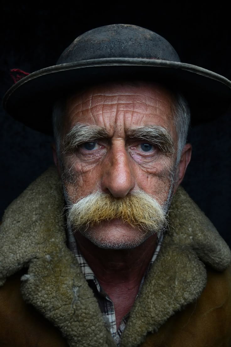 a closeup portrait of an elderly man with a striking mustache, wearing a hat and fur coat. the man has a weathered face with visible wrinkles, suggesting a life of experience. his expression is serious and intense, his eyes are fixed on the viewer. the lighting emphasizes the details of his face, adding to the depth of the image. the man is wearing a hat, which adds a traditional touch to the portrait.