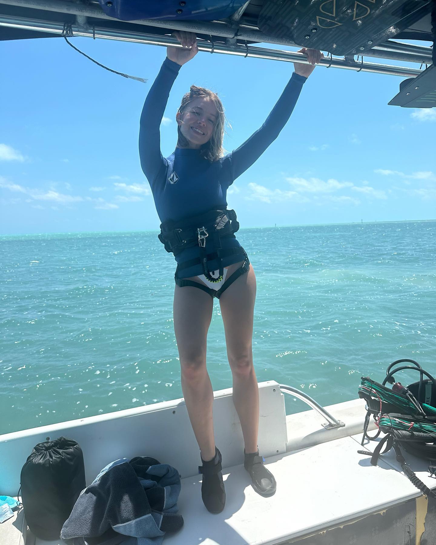 a young woman in a blue kitesurfing wetsuit and harness stands on a boat, holding onto a kiteboarding bar overhead.  the ocean is a calm turquoise blue under a bright, sunny sky.  she is smiling and appears happy and relaxed.  other kitesurfing equipment is visible on the boat. the overall scene suggests a fun, active vacation or day of watersports.