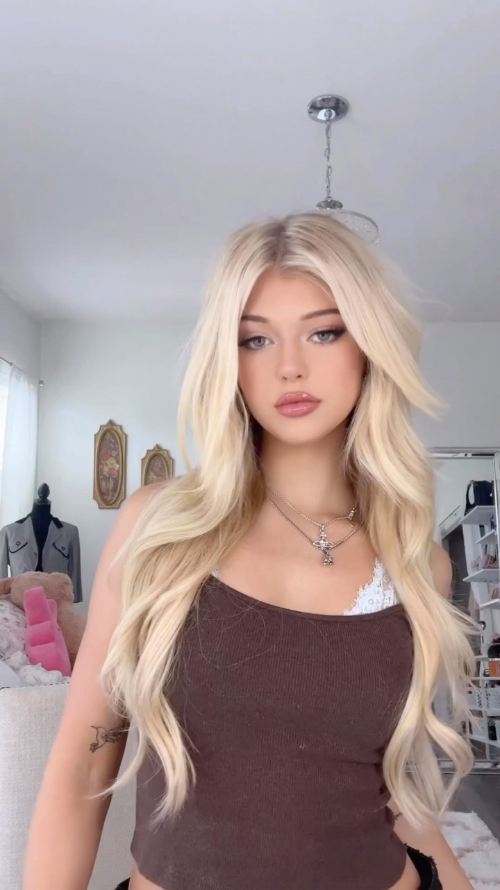 a portrait of loren gray with long blonde hair, wearing a brown tank top and a silver cross necklace, indoors. there is a light fixture above her head, and a mannequin is on the left side of the image. she has fair skin and lip filler. a glimpse of her tattoo can be seen on her left arm. the background appears to be a bedroom.
