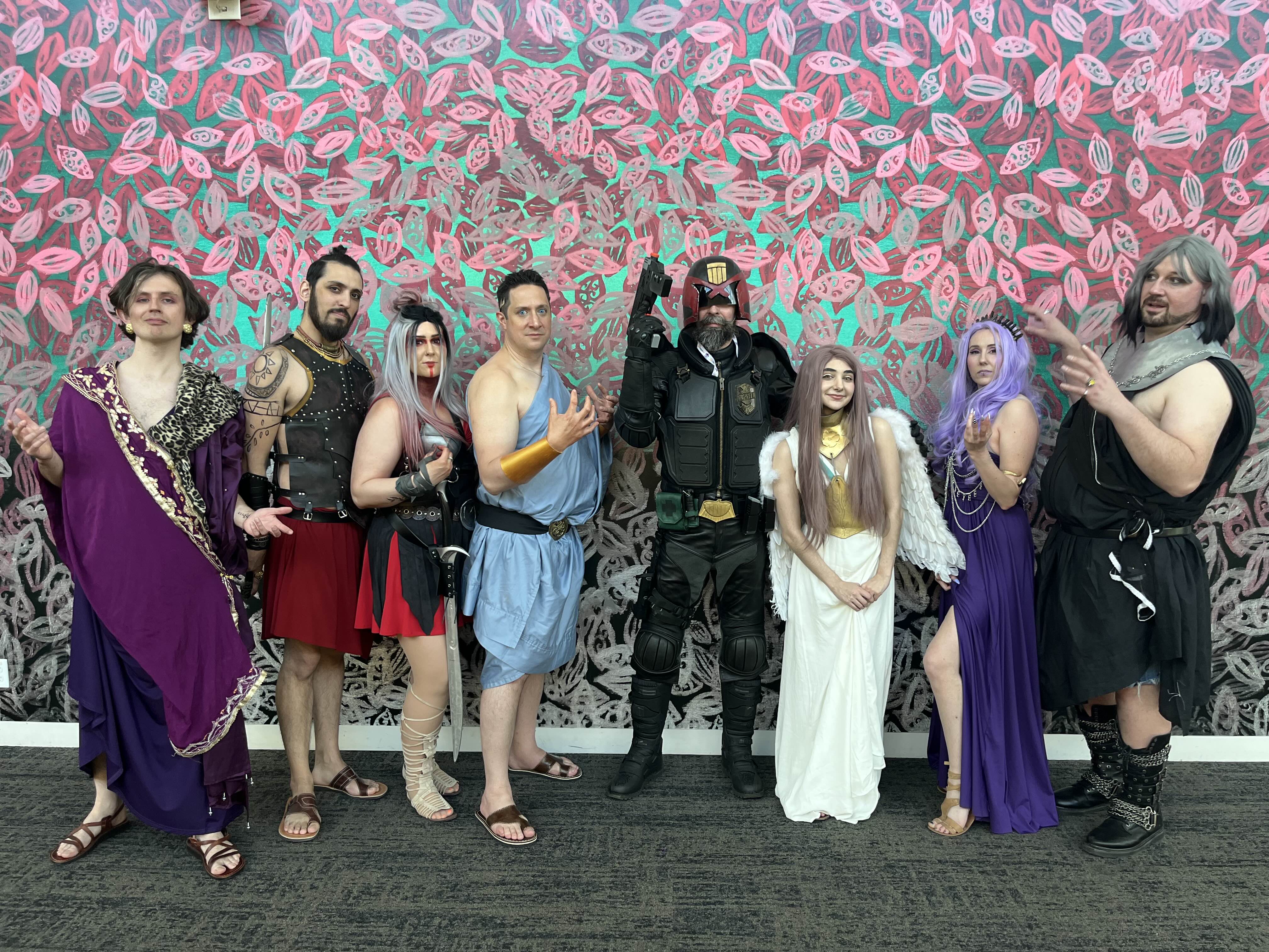 group of people in elaborate cosplay costumes pose for a photo in front of a vibrant, patterned wall. the costumes represent a mix of characters, including judge dredd (a scifi character), several individuals in attire reminiscent of ancient greek gods and goddesses, and an angel. the diverse group showcases creativity and enthusiasm for their chosen characters. the setting suggests a convention or similar event.