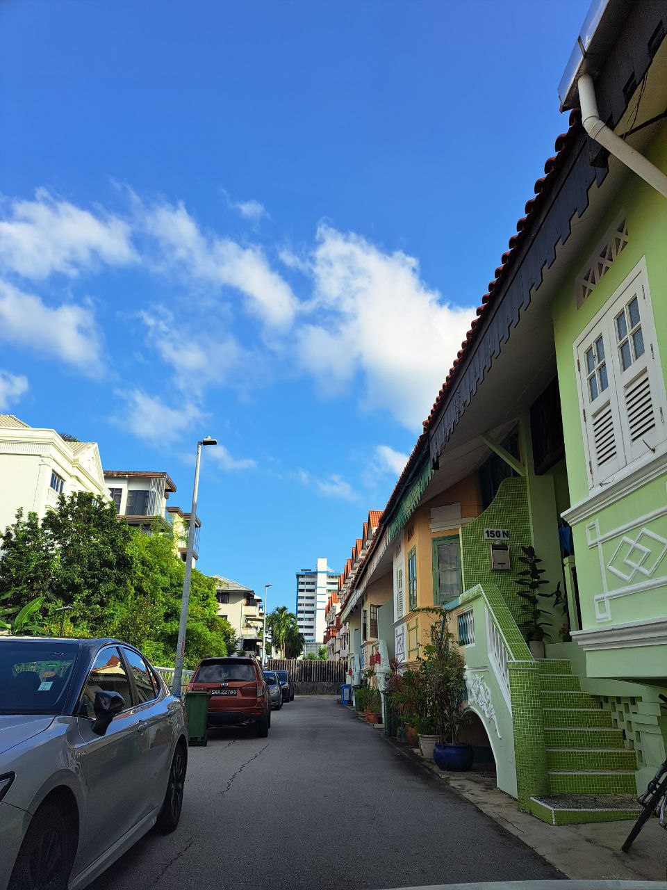 a narrow street lined with colorful houses, a few parked cars, some greenery, and a bright blue sky with fluffy clouds.  the perspective of the image draws the viewer's eye down the street towards the horizon.