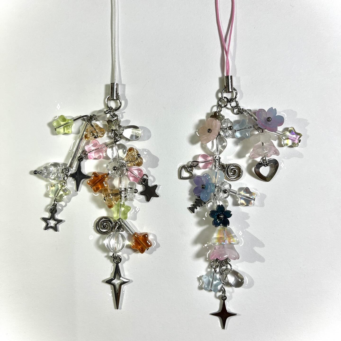 the image shows two beaded phone charms. one charm has a white cord and is made up of beads in a variety of colors including pink, orange, green, and clear, with star and crossshaped charms. the other charm has a pink cord and features beads in shades of pink, blue, and clear, along with flower, heart, fish, and crossshaped charms.