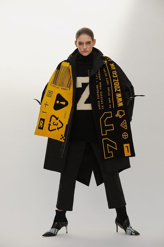a young woman stands in a studio, posing in a black coat and black pants, paired with a yellow scarf. the scarf is a statement piece with a black and yellow design. the model is wearing stylish black boots and looking confidently towards the camera. her attire embodies a mix of casual and chic with a modern twist.