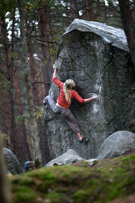 a woman in athletic wear is bouldering on a large rock formation in a forest. the image is taken from an eyelevel perspective, capturing the climber in midascent. the forest setting provides a natural backdrop, with trees, moss, and soft, diffused lighting.