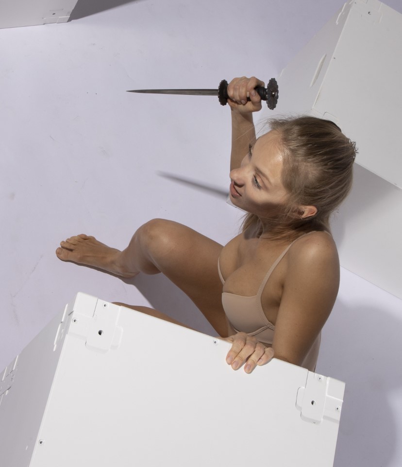 a woman in underwear holds a knife while sitting amongst white boxes.