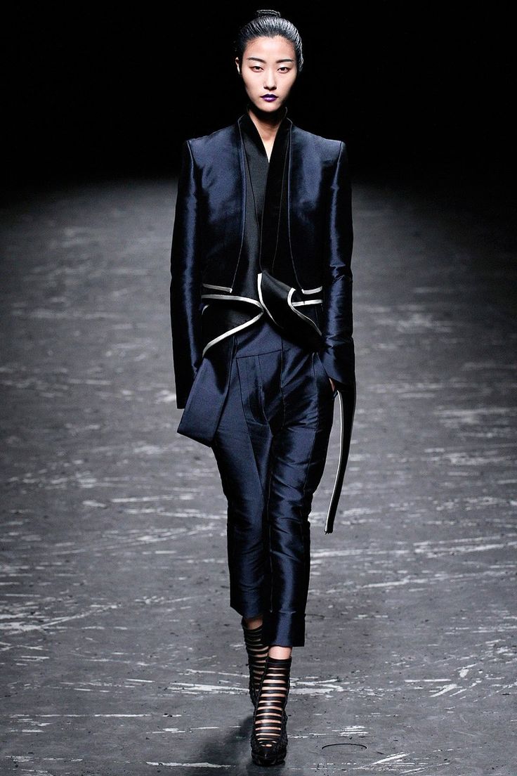 a fashion model walks the runway in a black and blue outfit. the model is wearing a longsleeved navy blue jacket with a black trim and a white trim on the front. the model is wearing black trousers, and her hair is tied up. the model is wearing black highheeled boots that have straps. the model is posing and looking straight ahead at the camera.