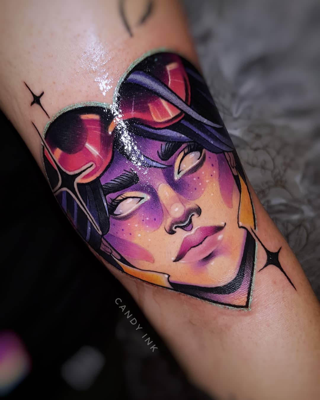 a closeup shot of a colorful tattoo on someone's inner arm. the tattoo depicts a female video game character with purple hair and white eyes, wearing pink goggles on top of her head. her face is framed by a heart shape. the tattoo is done in a neotraditional style with bold lines and shading. the artist's signature, "candy ink," is visible below the tattoo.
