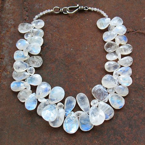 a moonstone necklace is laid out on a rusty metal surface. the necklace features teardropshaped moonstones of varying sizes, arranged in a cascading pattern. the stones are translucent and exhibit a soft, iridescent sheen, with hues of blue and white. a silver clasp secures the necklace. the rusty metal background provides a contrasting backdrop to the delicate and shimmering stones.