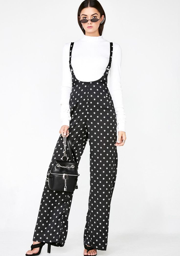 a woman stands in front of a white background wearing a white turtleneck and black overalls with a white star pattern. she is holding a small black backpack with a chain and wearing black heeled sandals and sunglasses.