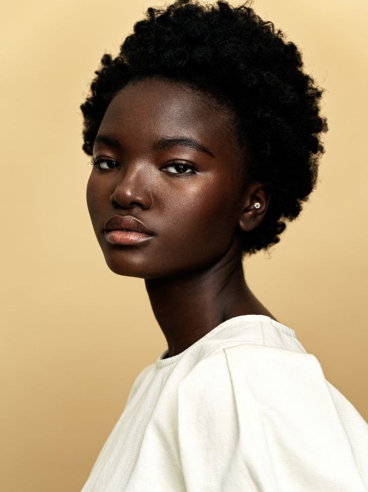 a portrait of a beautiful darkskinned woman with short, natural hair. she has a subtle earring and is wearing a white top. the background is a neutral beige. the photo is a shoulder shot focusing on her face and upper body.