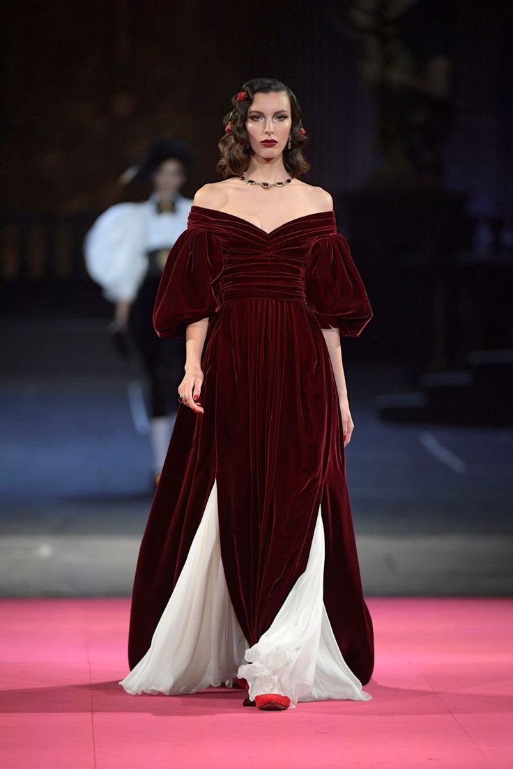 a model walks down a pink runway wearing a stunning burgundy velvet gown with offtheshoulder puff sleeves.  the dress features a white underlay that peeks out from the side slits.  the model's hair is styled in a classic hollywood wave with small red flowers adorning it. she wears a simple necklace and red shoes.  the background is slightly blurred, but shows what appears to be a dark, formal setting.  the overall impression is one of high fashion and elegance.