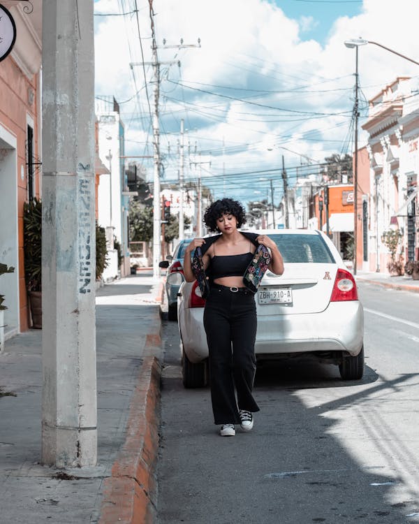 a young woman with dark hair walks down a city street. she is wearing black pants, a black crop top, and a black and floral jacket. she is carrying her jacket over her shoulder. she is walking past parked cars. the street is lined with buildings.